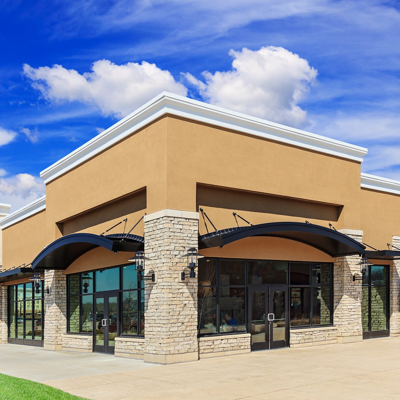 New commercial development featuring a street view of a Strip Mall with green grass, sidewalk and patio space. Blue sky and clouds are in the background.