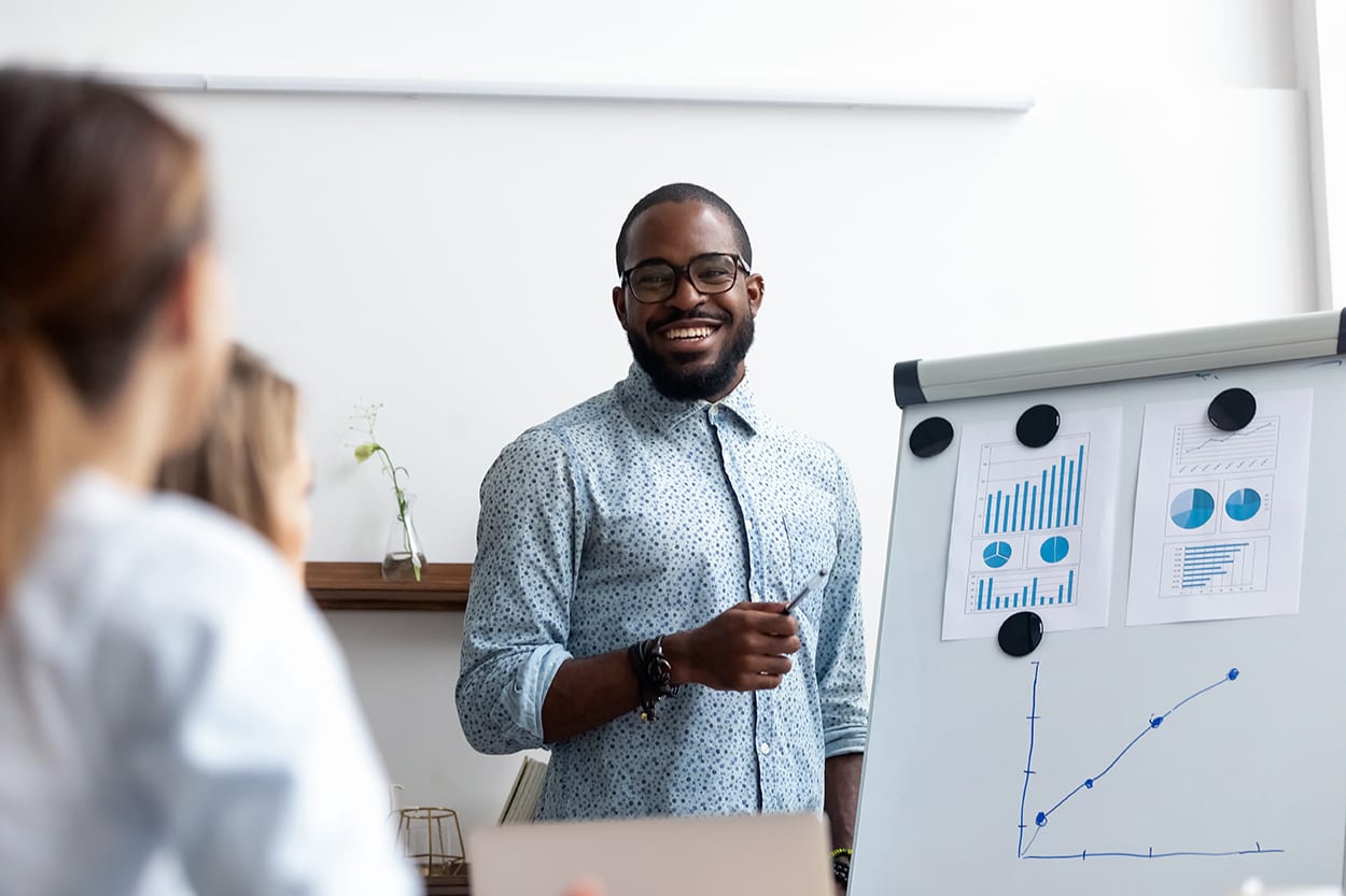 coach presents for company members financial report standing near whiteboard where shown sales statistics in graphs. Confident company owner teach interns acquaints with nuances of the market