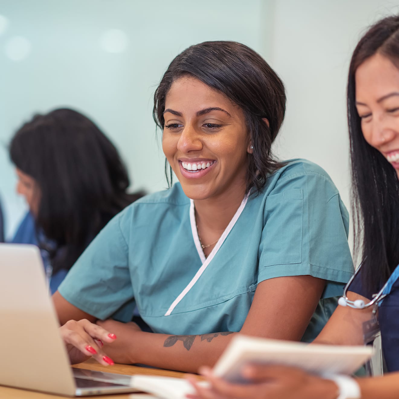 A multi-ethnic group of female nursing students are attending class together. They are seated at a long table in a classroom. The individuals are working in pairs on a project. The two individuals seated closest to the camera are having a discussion while using a laptop computer.