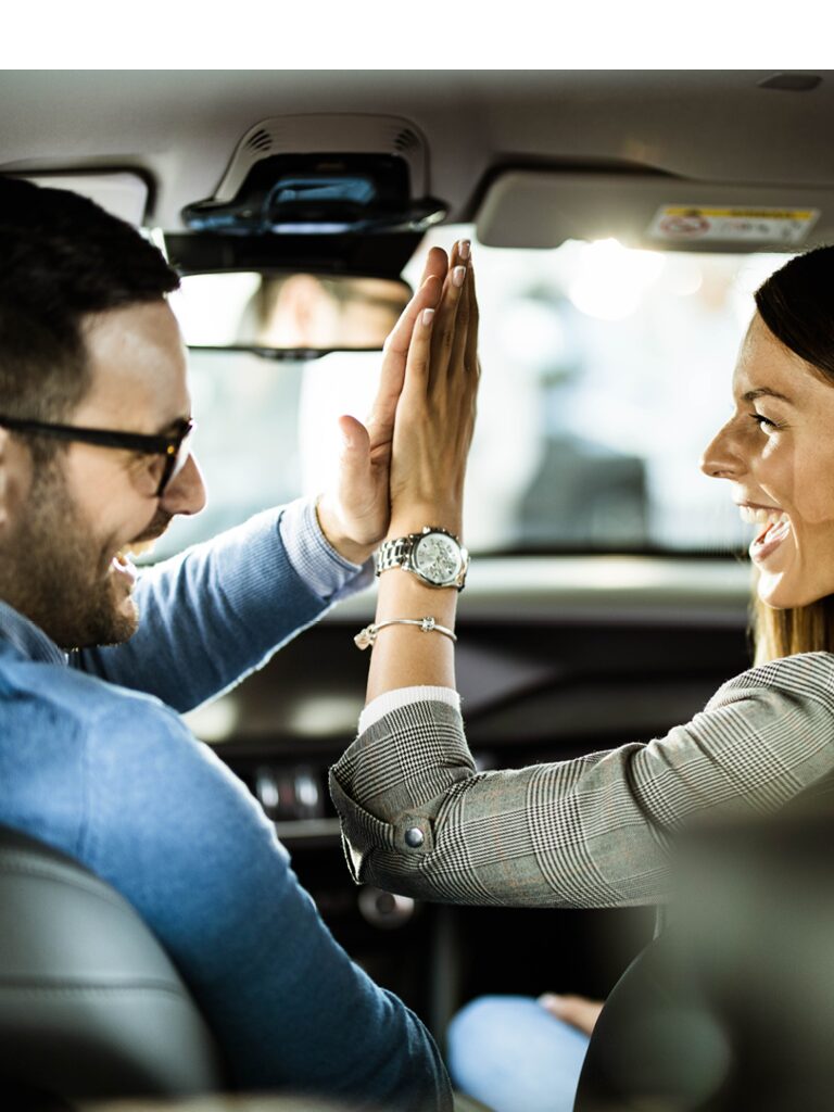 Image of a man and a woman in the front seat of a car. They are giving each other a high five and they are laughing.