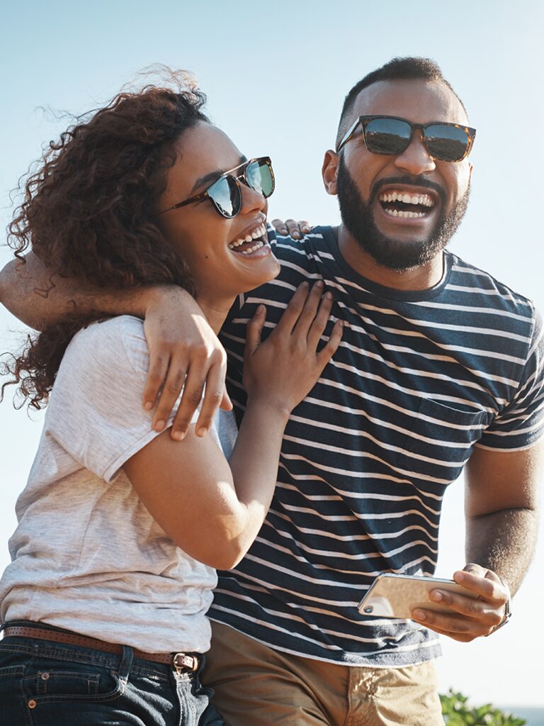 A man and a woman are smiling and laughing. He has his arm wrapped around her shoulder. They are outside on a sunny day.